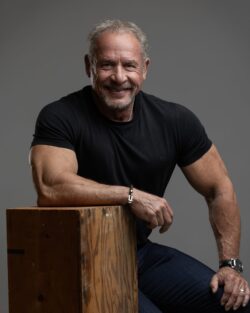 Studio portrait of a fitness coach leaning on a wooden box, black t-shirt, gray backdrop, Carmel, Indiana